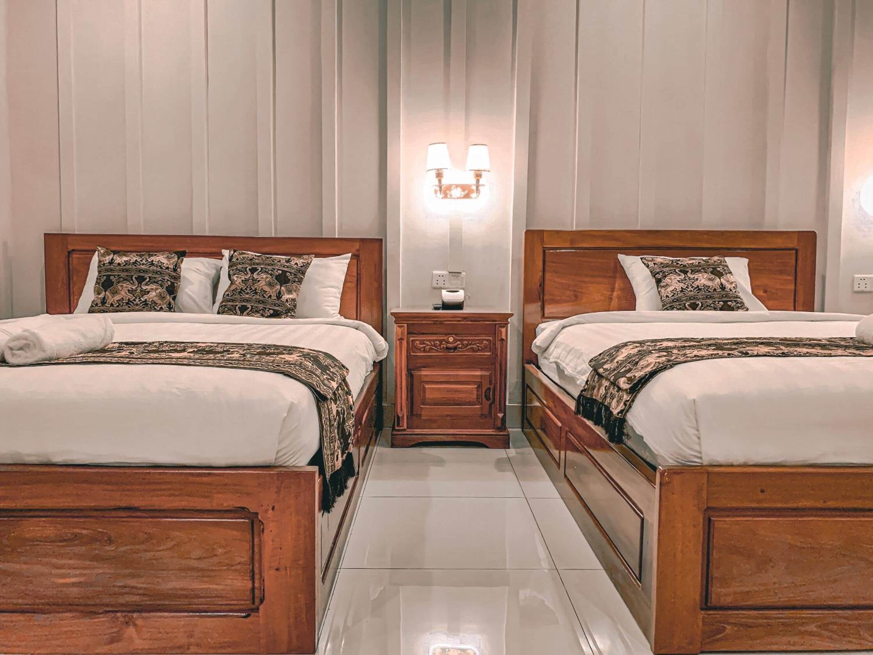 A hotel room with two wooden beds, each with patterned bedding and pillows. There is a nightstand with a lamp between the beds.