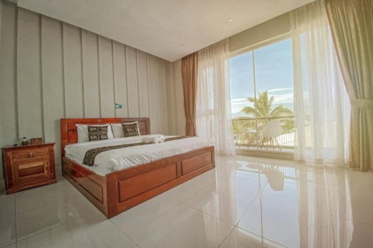 A hotel room with a large bed, a nightstand, a window with curtains, and a view of palm trees.