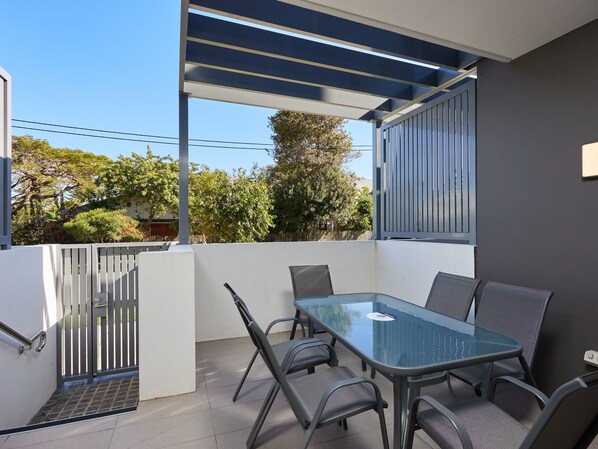 A balcony with a glass table and chairs, a metal gate, and a view of trees and buildings. - Ruby by Serain Residences (Surfers Paradise)
