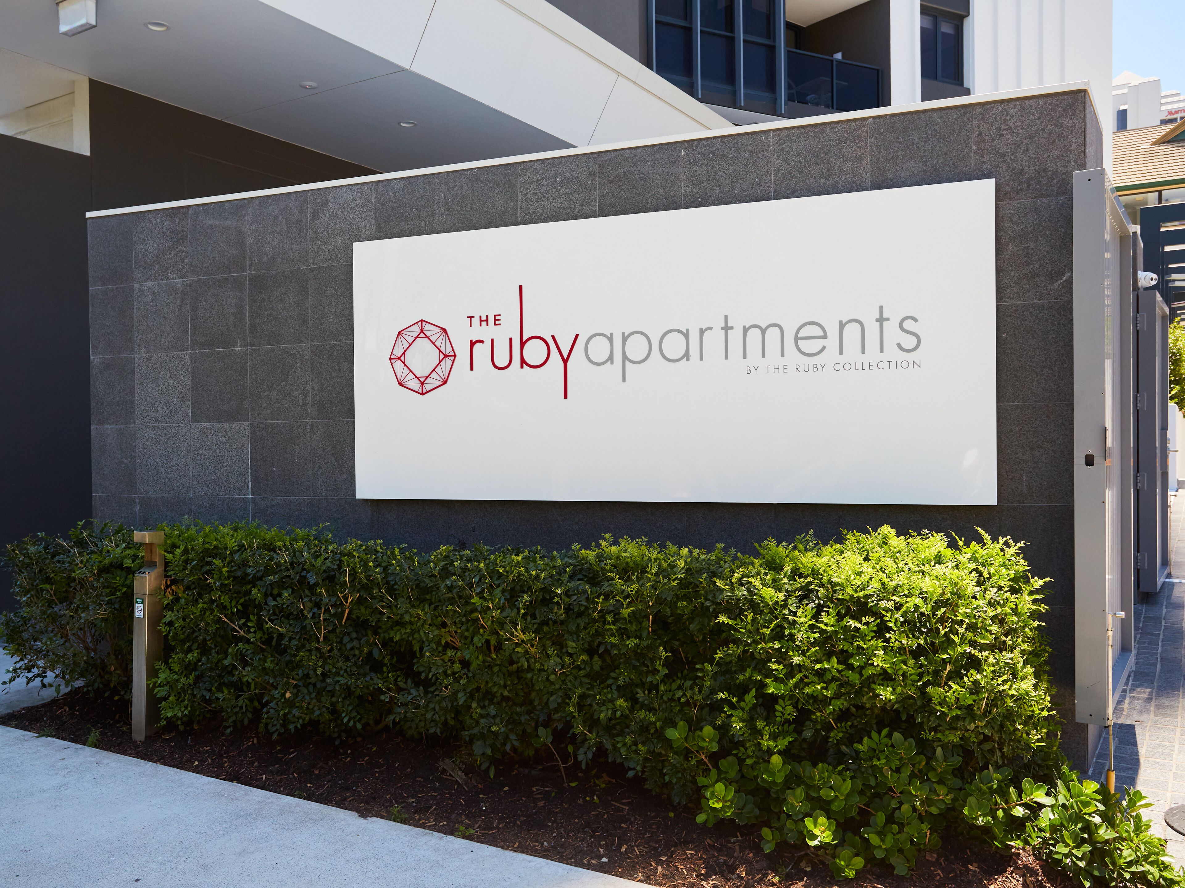 A sign for "The Ruby Apartments" with a geometric logo, set against a modern building facade with grey stone cladding and a green hedge in front.