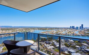 A balcony with wicker furniture overlooking a river and cityscape. - Ruby by Serain Residences (Surfers Paradise)