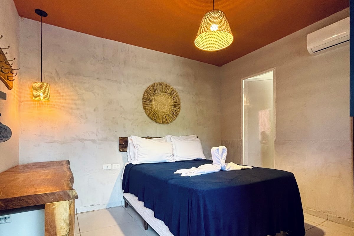 Un dormitorio con una cama, una mesita de noche de madera, una lámpara colgante y un aire acondicionado de pared.