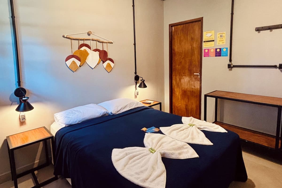Habitación de hotel con cama, mesitas de noche, escritorio, lámpara colgante y puerta de madera.