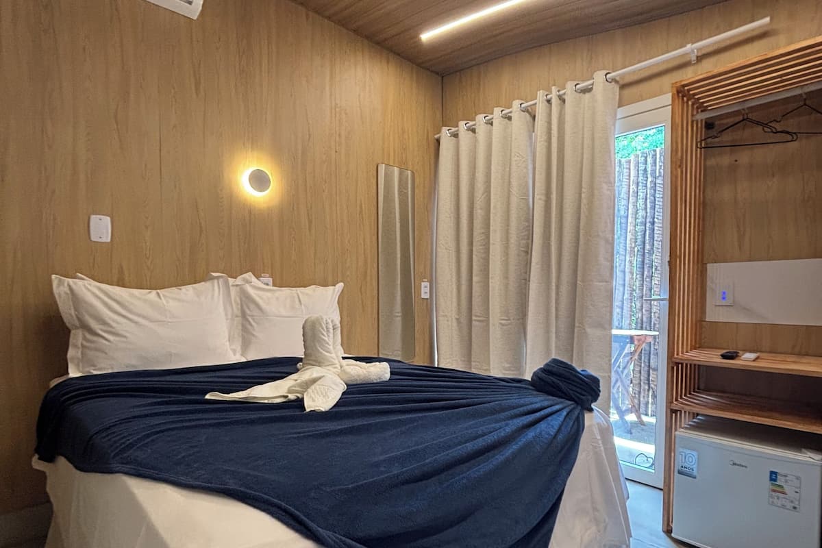 Un dormitorio con cama, mesita de noche, lámpara colgante, ventana con cortinas y armario.