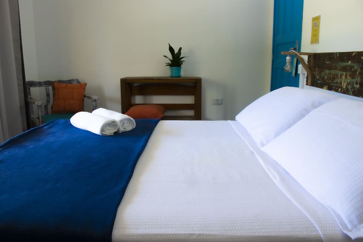Una cama bien tendida con sábanas blancas, un edredón azul y dos toallas enrolladas. Hay una mesita de noche de madera con una planta y una lámpara.
