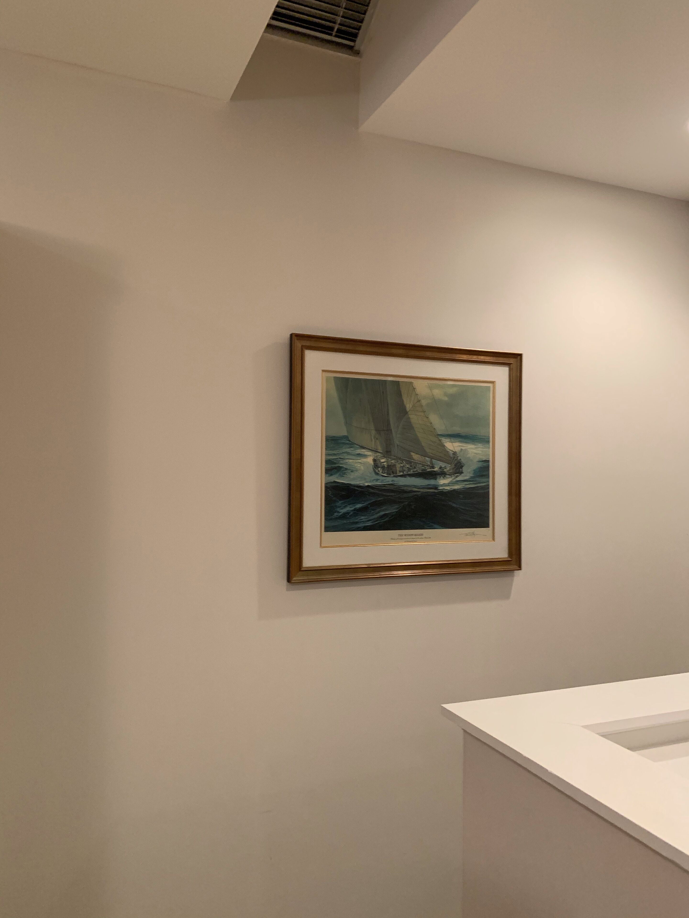 Un cuadro enmarco de un velero en un mar tormentoso, colgado en una pared lisa en una habitación con un lavabo blanco.