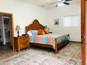 A bedroom with a wooden bed, a nightstand, a bedside table, a floor lamp, a patterned rug, a ceiling fan, and a window with blinds.