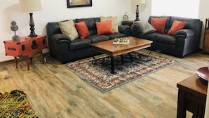 Un salon avec deux canapés noirs, une table basse en bois, un tapis à motifs, une valise rouge et un tableau encadré accroché au mur.