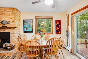 Enjoy meals in this cozy dining area with a large table and six chairs, surrounded by a stone fireplace and a window with a view of the outdoors.