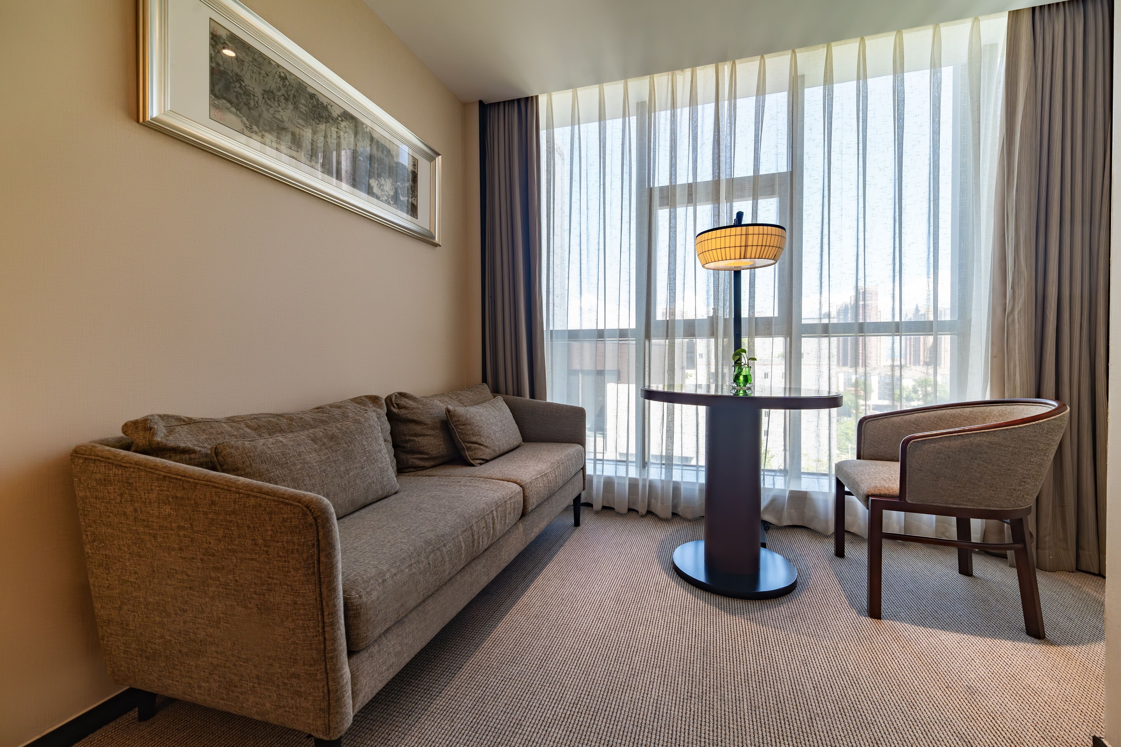 Una habitación de hotel moderna con sofá, una silla, una mesa con lámpara y un amplio ventanal con cortinas traslúcidas.