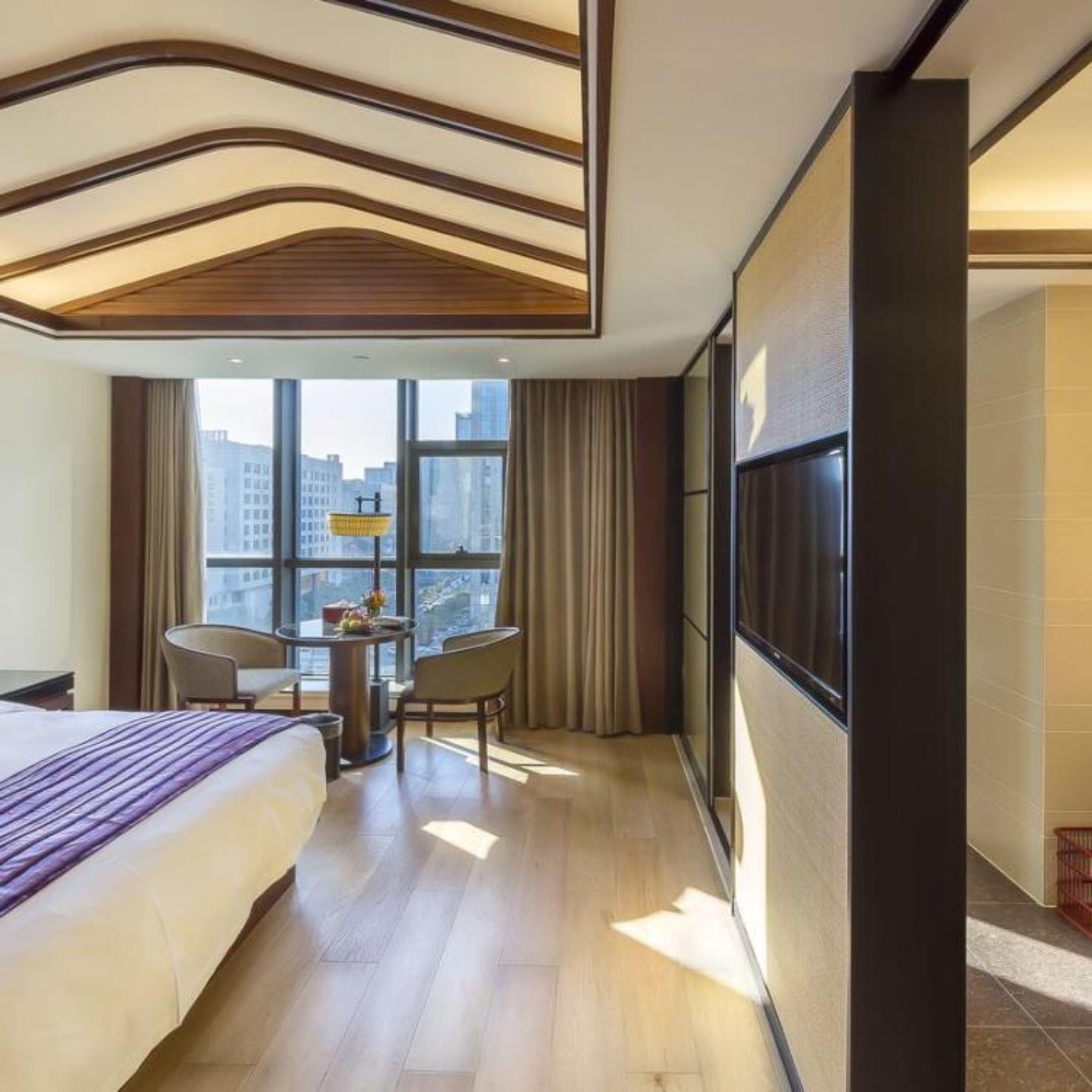 Een hotelkamer met een groot bed, een bureau met twee stoelen, een televisie aan de muur en uitzicht op de stad door grote ramen.