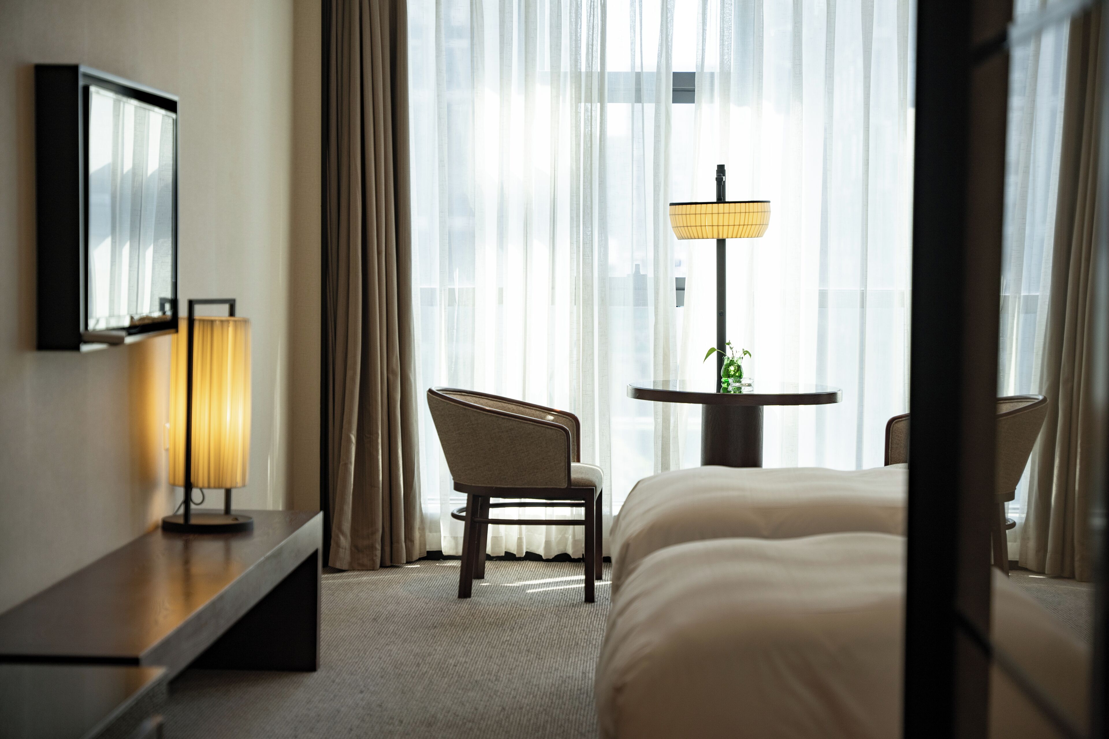 A hotel room with a bed, a desk, a chair, a table with a lamp, and a window with curtains.
