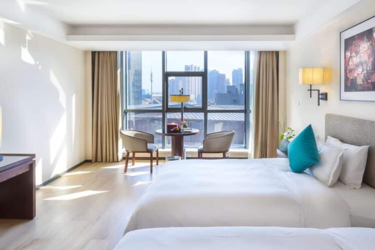 A hotel room with a large bed, a desk, a chair, and a view of the cityscape through large windows.