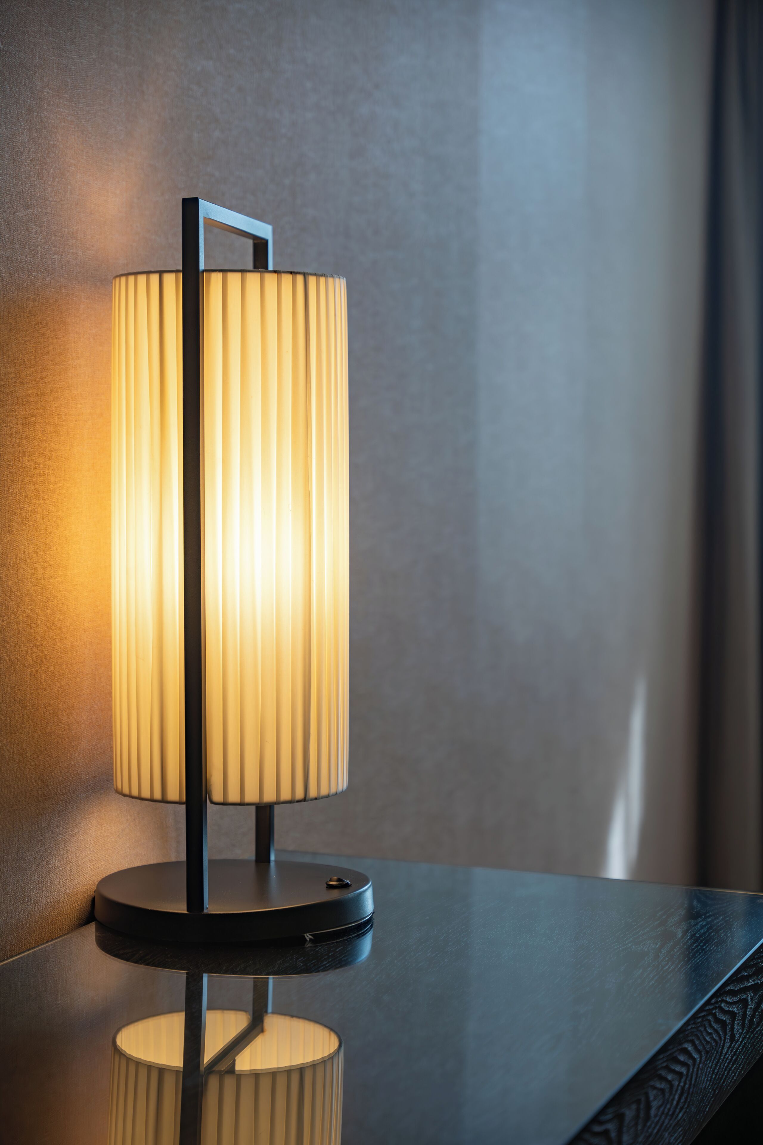 A modern table lamp with a cylindrical shade and a rectangular base, placed on a reflective surface.