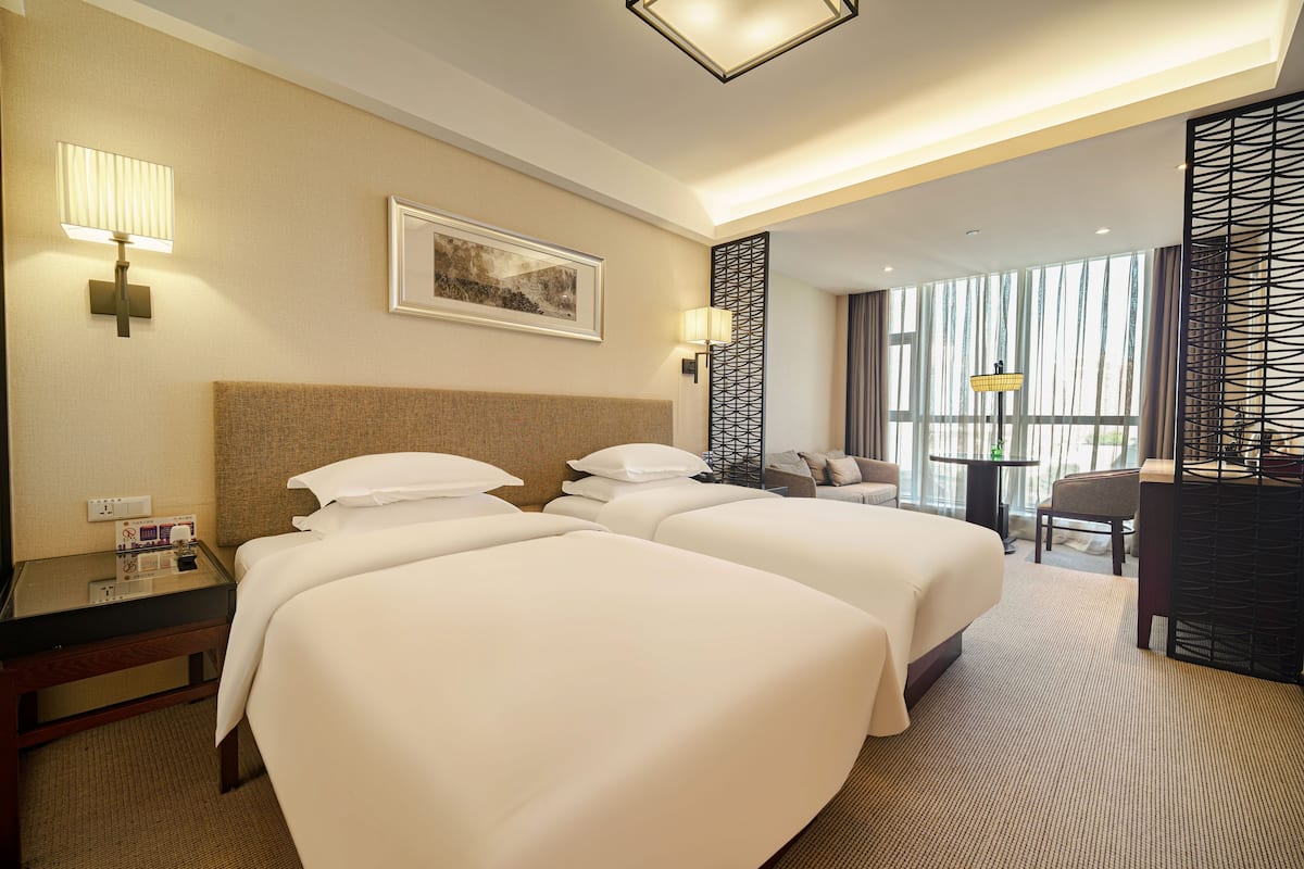 A hotel room with two beds, a desk with a lamp, a chair, and a large window with curtains.
