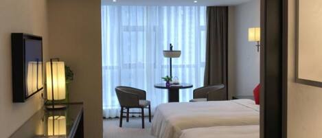A hotel room with two beds, a desk with a lamp, a chair, a small table with a plant, and a window with sheer curtains.