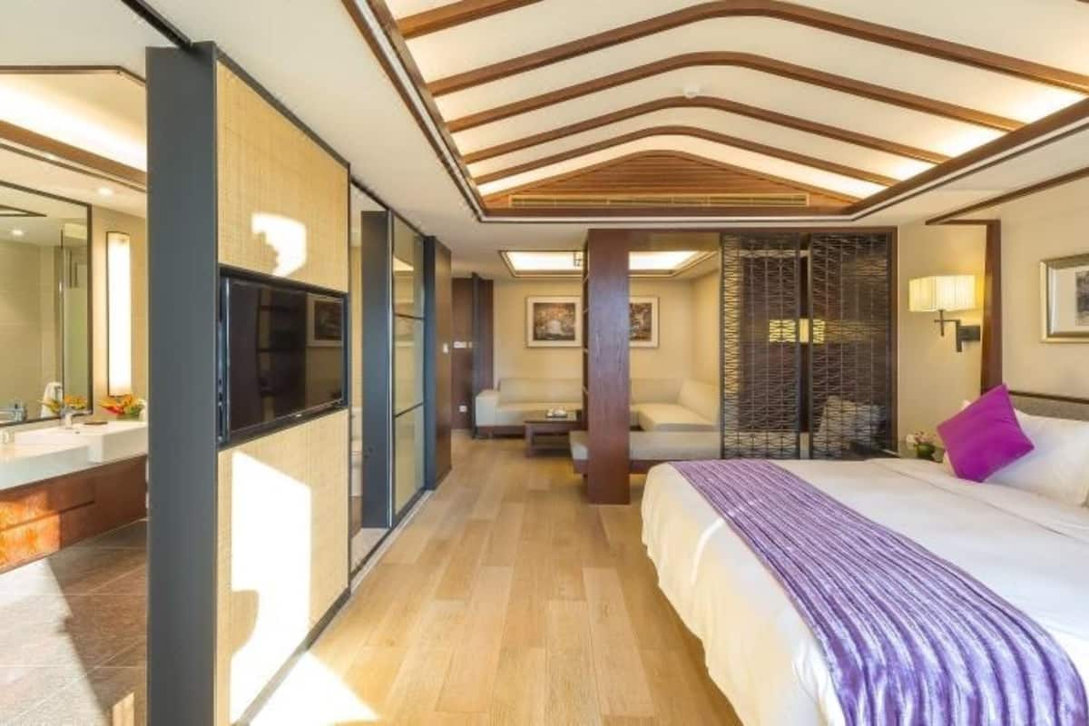 A modern hotel room with a large bed, a television, a bathroom with a mirror, and a wooden ceiling.