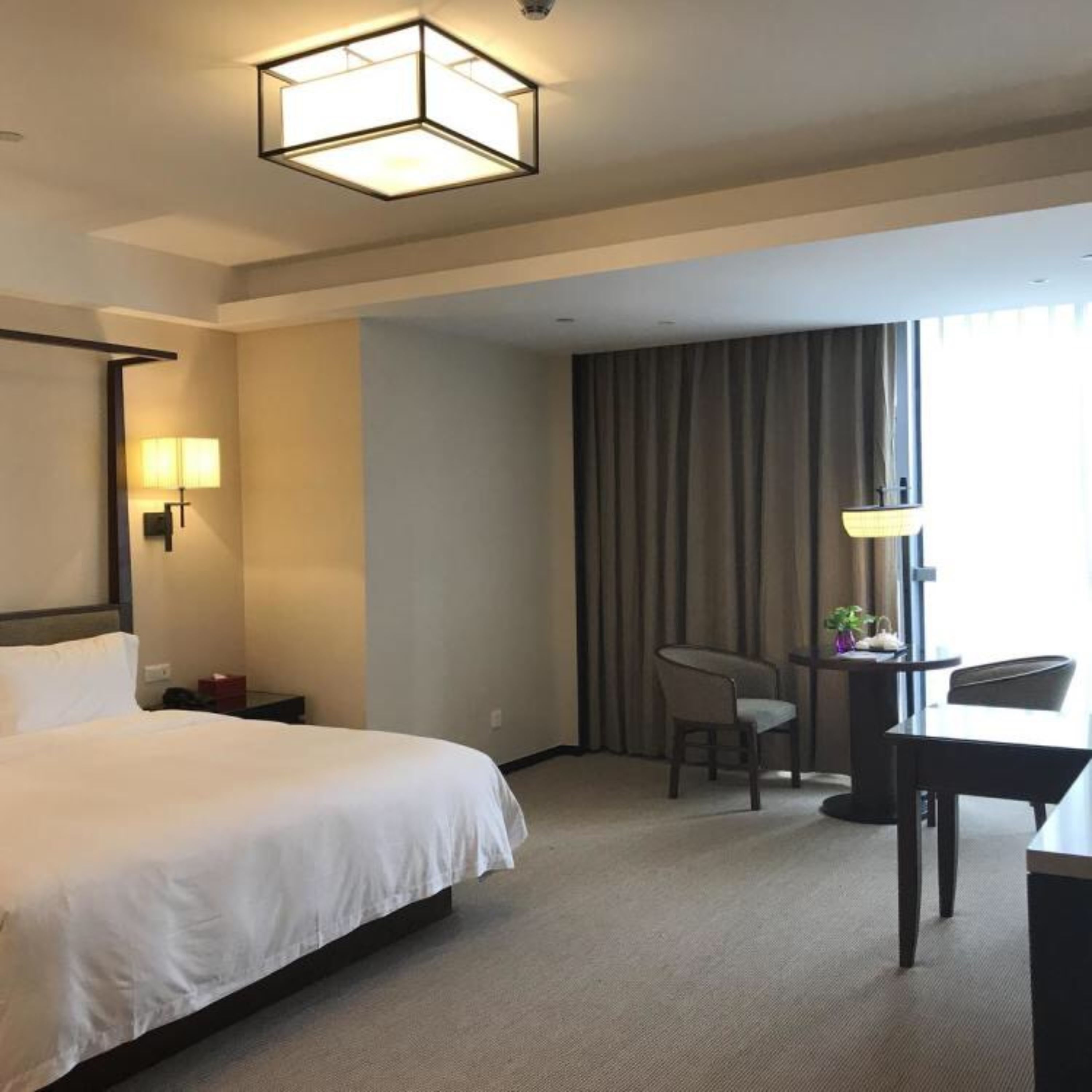Habitación de hotel con una cama grande, un escritorio, una silla, una lámpara de escritorio, una lámpara de pared y un ventanal con cortinas.