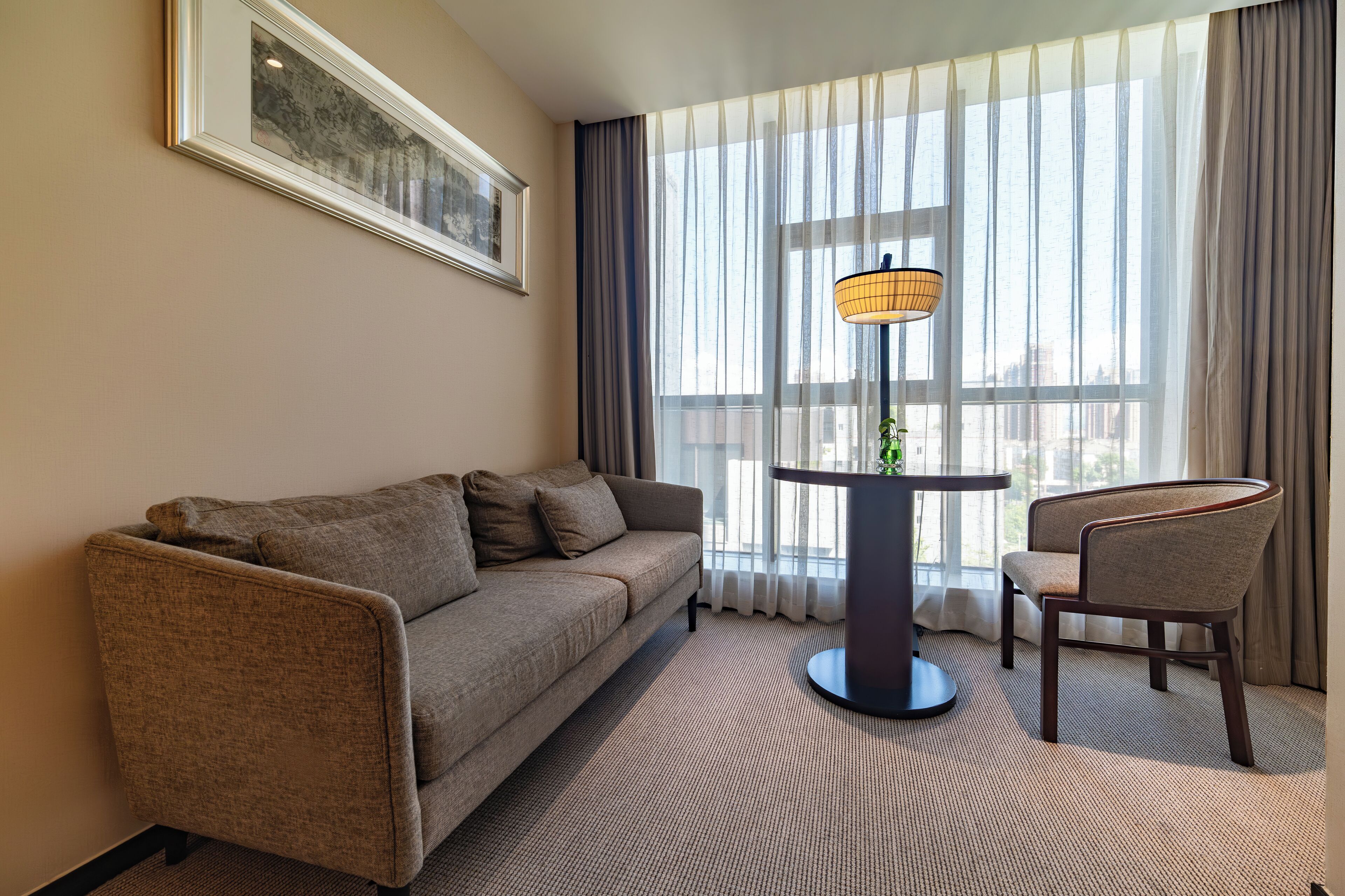 Habitación de hotel con sofá, una silla, una mesita con lámpara y un ventanal con cortinas translúcidas.