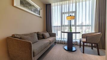 Habitación de hotel con sofá, una silla, una mesita con lámpara y un ventanal con cortinas translúcidas.