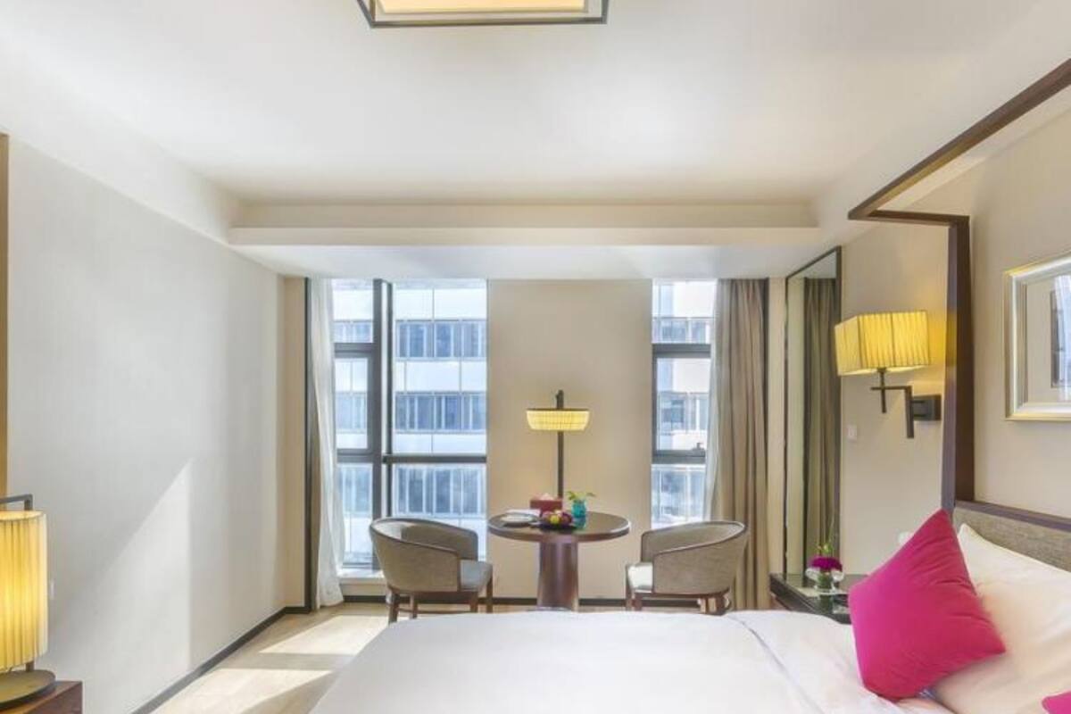 A hotel room with a large bed, two chairs, a small table, and a window with a view of buildings.