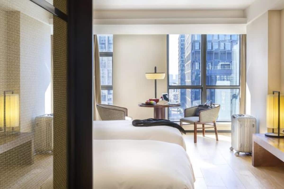 A hotel room with a large bed, a desk with a lamp, a chair, and a view of the city through large windows.