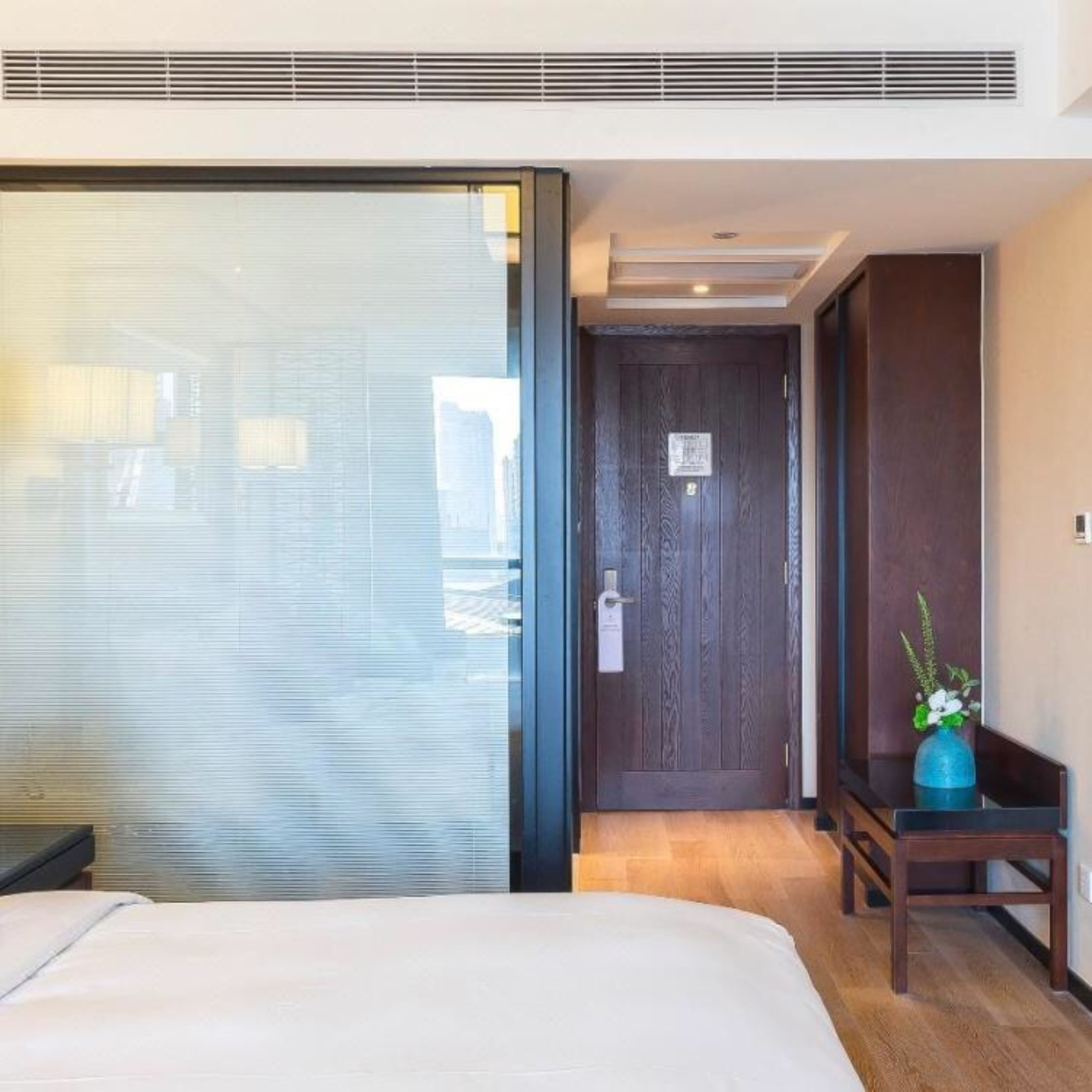 A hotel room with a large bed, a sliding glass door, a dark wooden door, and a small table with a vase of flowers.