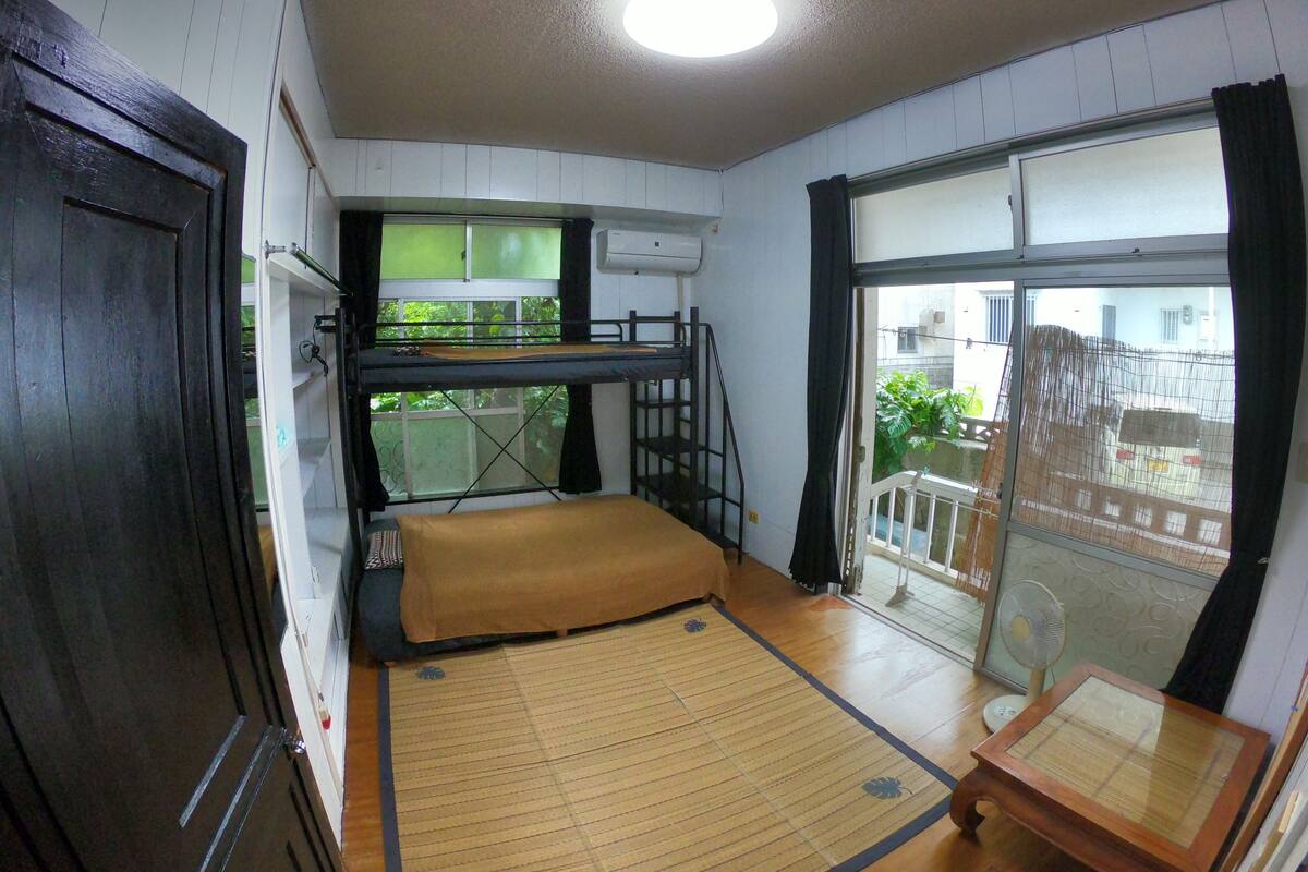 A compact room with a bunk bed, a tatami mat, a small table, and a sliding door leading to a balcony.