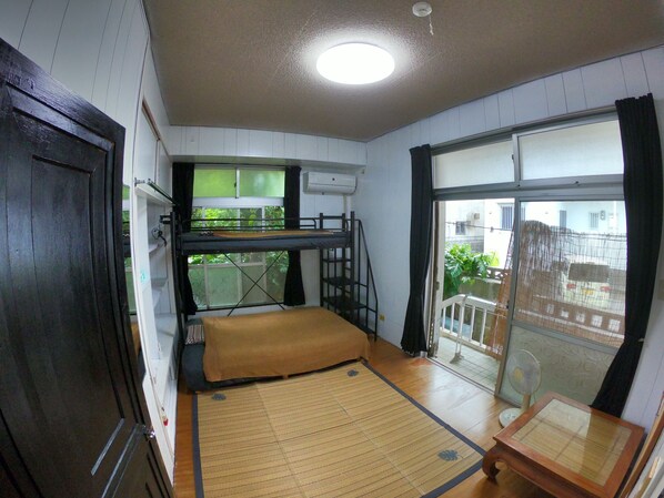 A compact room with a bunk bed, a tatami mat, a small table, and a sliding door leading to a balcony.