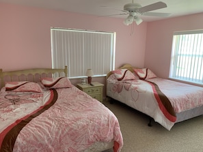 Quarto com duas camas, um ventilador de teto, janela com persianas e mesa de cabeceira com abajur.
