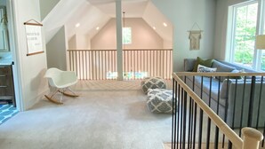 A spacious room with a high ceiling, a sloped roof, a white rocking chair, a sofa with cushions, and a view of the outdoors through large windows.