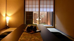 Une pièce de style japonais traditionnel, avec un sol recouvert de tatamis, une petite table sur laquelle est disposé un service à thé, des portes coulissantes en papier de riz (shoji) et une vue sur une clôture en bois et un arbre.