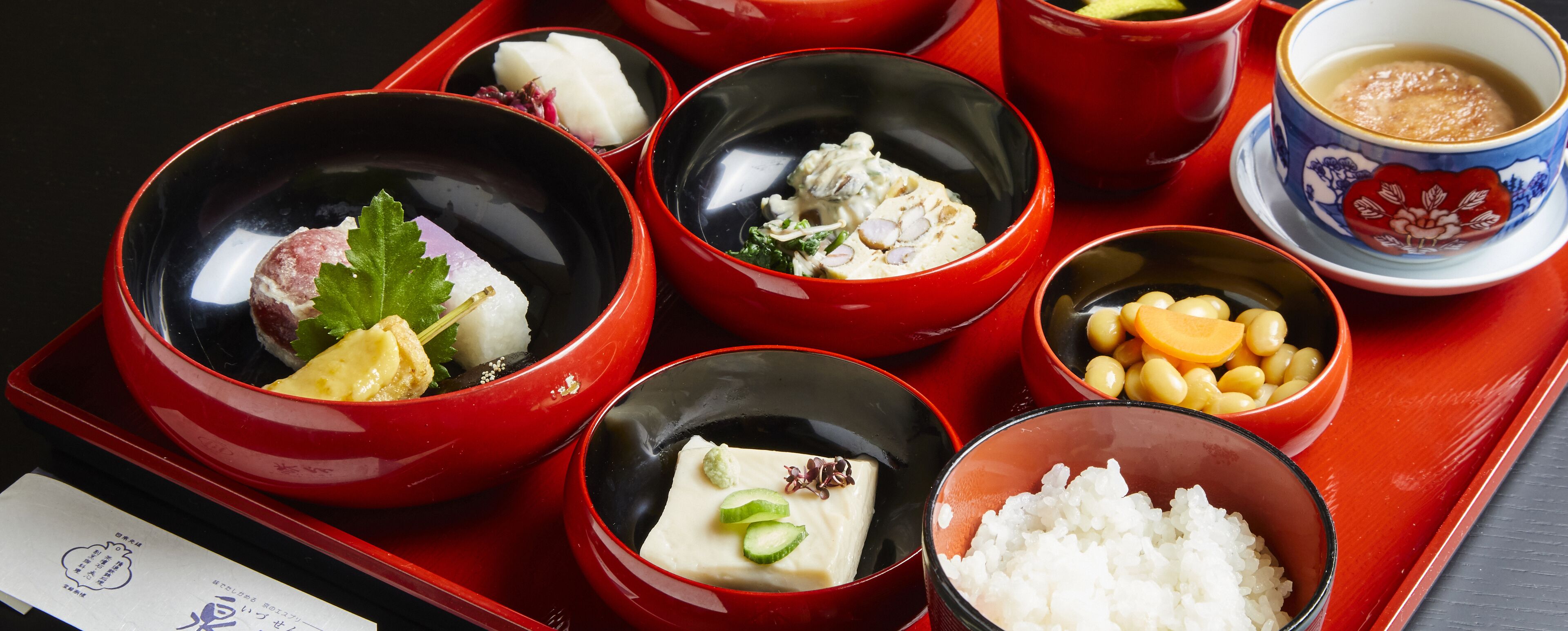 A traditional Japanese meal with various dishes in red and black bowls, including rice, fish, and pickled vegetables, served on a red tray.