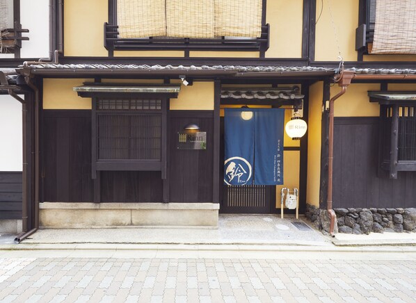 A traditional Japanese building with a dark wooden entrance, a blue awning with a logo, and a stone lantern outside. - Rinn Shijo Takasegawa North (Kyoto)