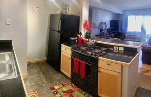 A compact kitchen with a stove, oven, and refrigerator. There is a small island with a kettle and a red towel hanging on the oven. A clock is visible on the wall.