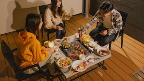 Un groupe de personnes est réuni autour d’une table en bois, à l’extérieur, et déguste divers plats préparés sur un grill.