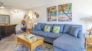 A living room with a blue sofa, a glass coffee table, and a kitchen area with white cabinets.