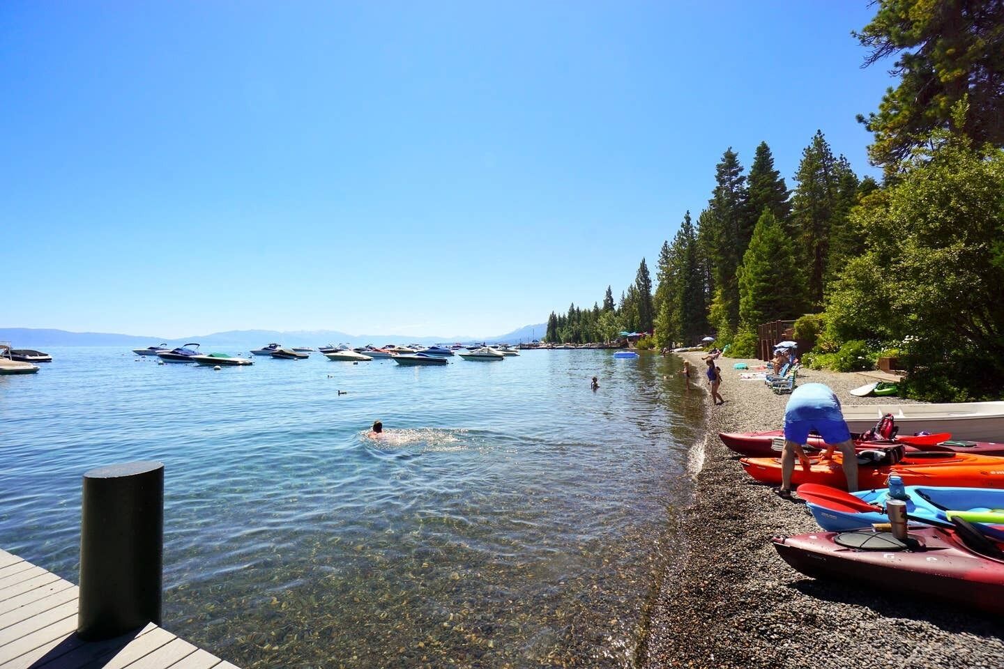 Escena junto al lago con kayak y botes en el agua, un muelle de madera y una playa de piedras.