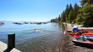 Une scène au bord d’un lac, avec des kayaks et des bateaux sur l’eau, une jetée en bois et une plage de galets.