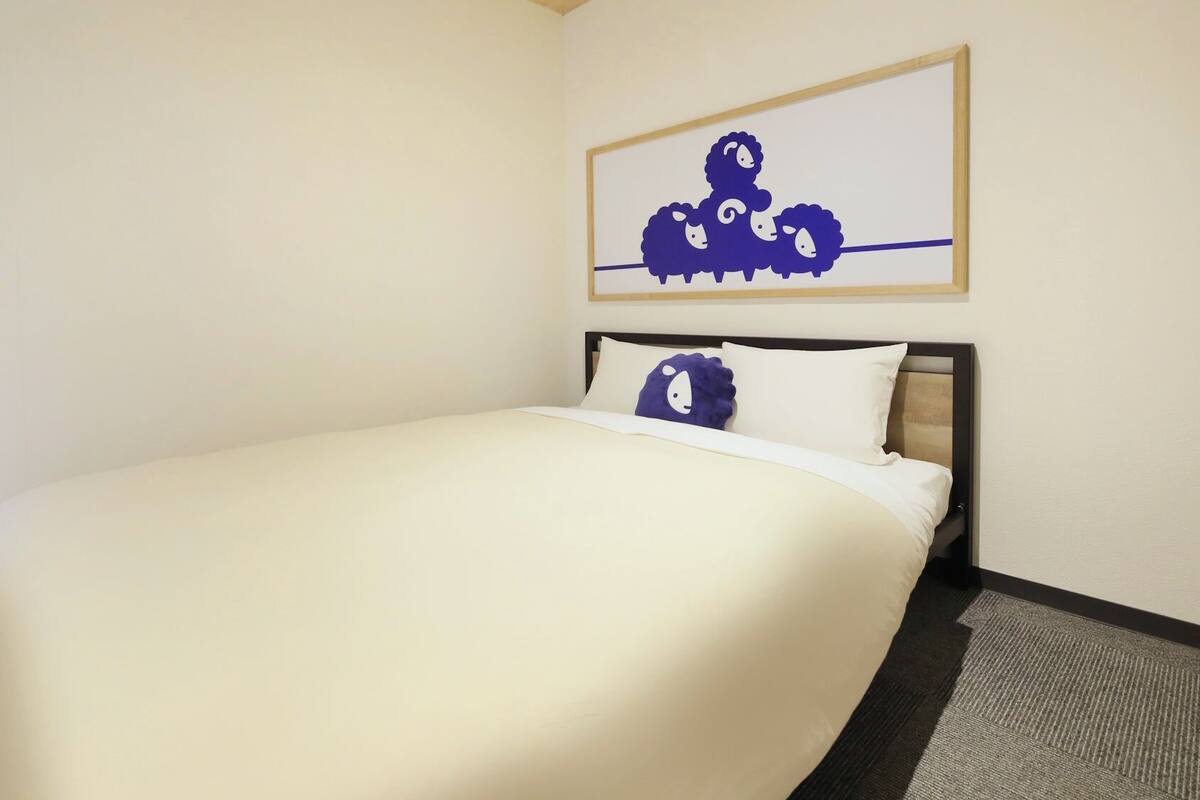 A hotel room with a bed, a headboard, a wall-mounted artwork of sheep, and a carpeted floor.