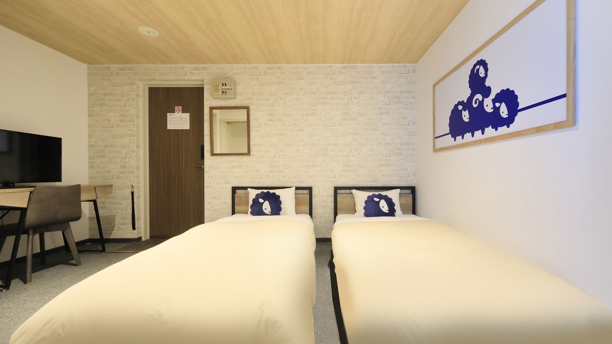 A hotel room with two beds, a desk with a chair, a television, a brick-patterned wall, and a framed picture on the wall.