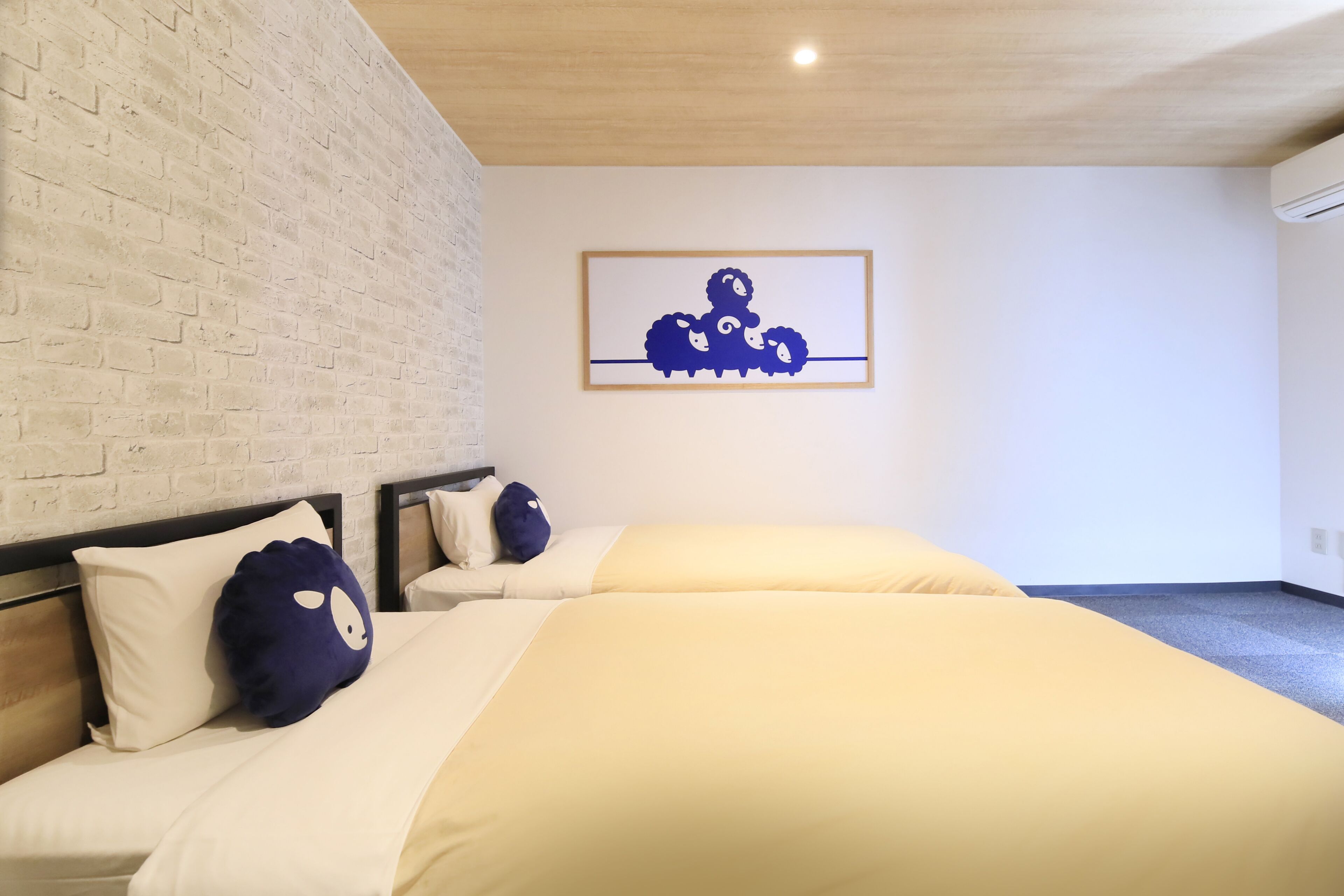 Habitación de hotel con pared de ladrillo, cama con ropa de cama color crema y una almohada azul, y un cuadro con ovejas en la pared.