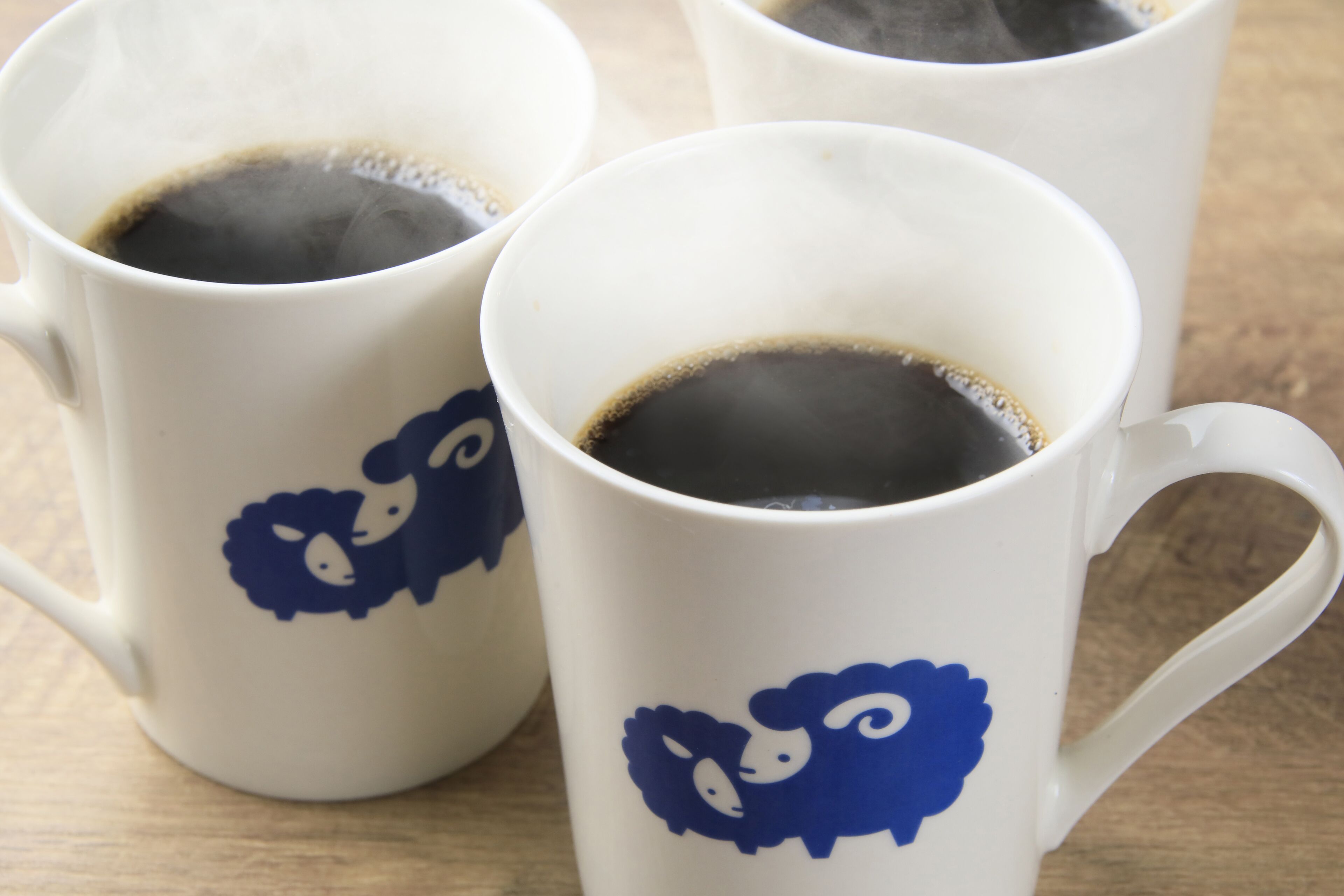 Three white ceramic mugs with blue sheep designs, each containing a dark liquid, likely coffee, on a wooden surface.