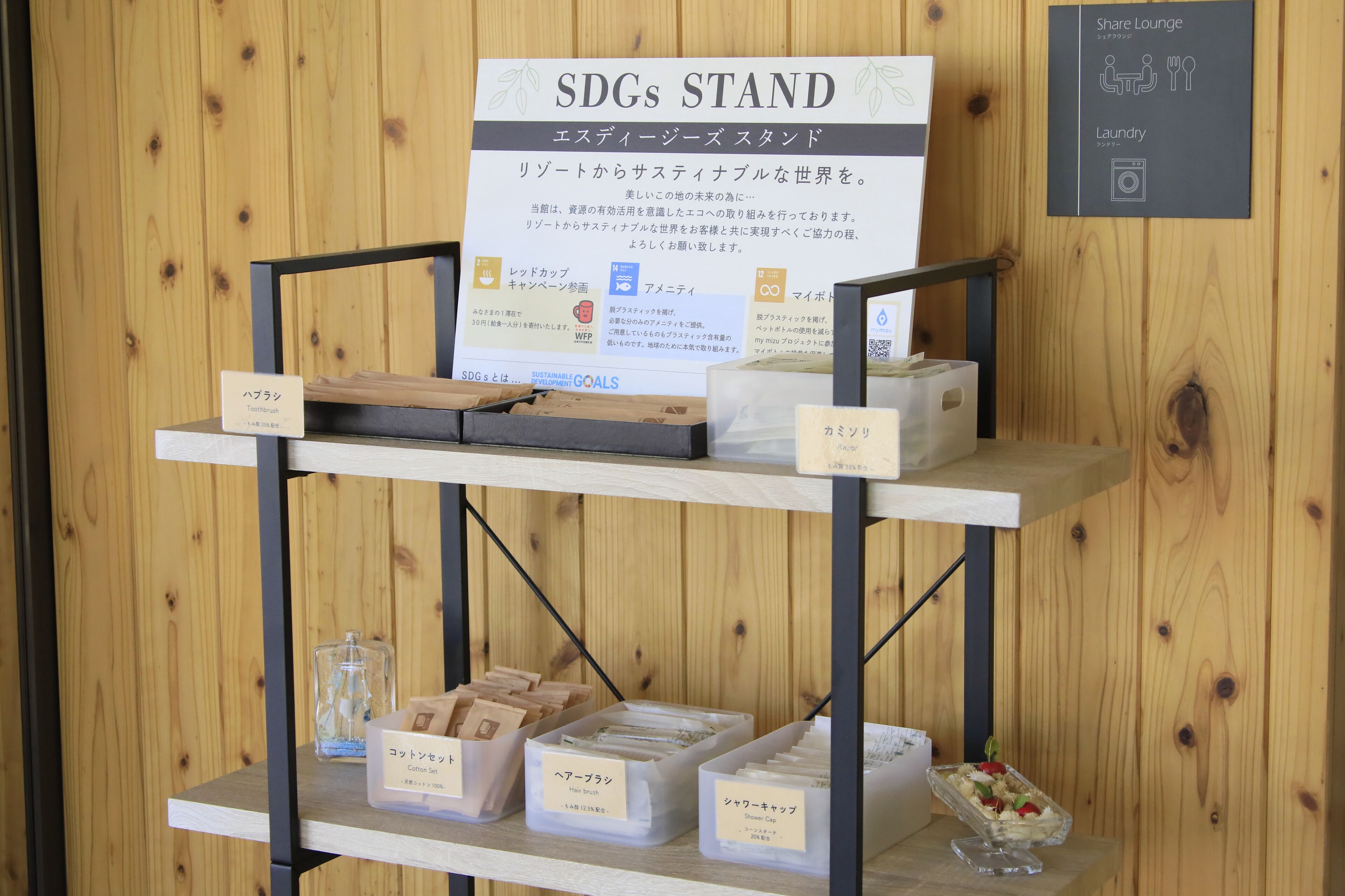 Una pared de madera con un letrero que dice "SDGs STAND" y una repisa con varios objetos, incluyendo cajas etiquetadas "cepillo para el cabello" y "gorro de ducha".