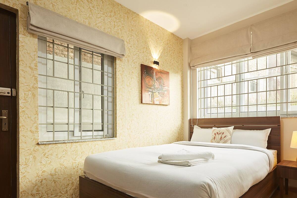 Ein Hotelzimmer mit einem Bett, zwei Jalousien an den Fenstern, einer Nachttischlampe und einem Bild an der Wand.