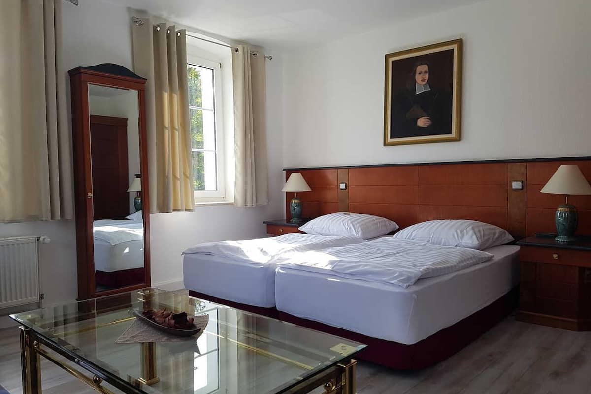 Ein Hotelzimmer mit einem Doppelbett, einem Glastisch, einem Holzkopfende, einem Nachttisch mit Lampe, einem Spiegel und einem fenster mit Vorhängen.