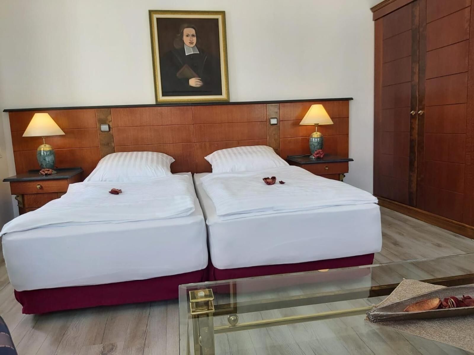 A hotel room with two beds, a wooden headboard, two bedside tables with lamps, a framed portrait on the wall, and a wooden wardrobe.
