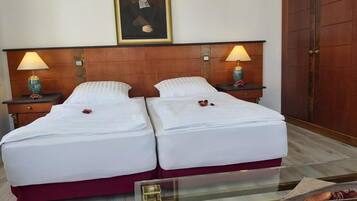 A hotel room with two beds, a wooden headboard, two bedside tables with lamps, a framed portrait on the wall, and a wooden wardrobe.
