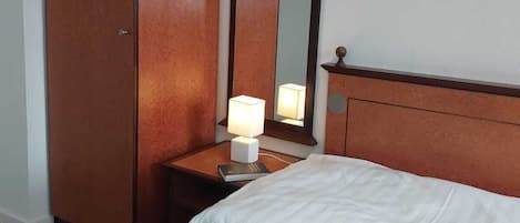 A hotel room with a bed, a wardrobe, a nightstand with a lamp, and a mirror.