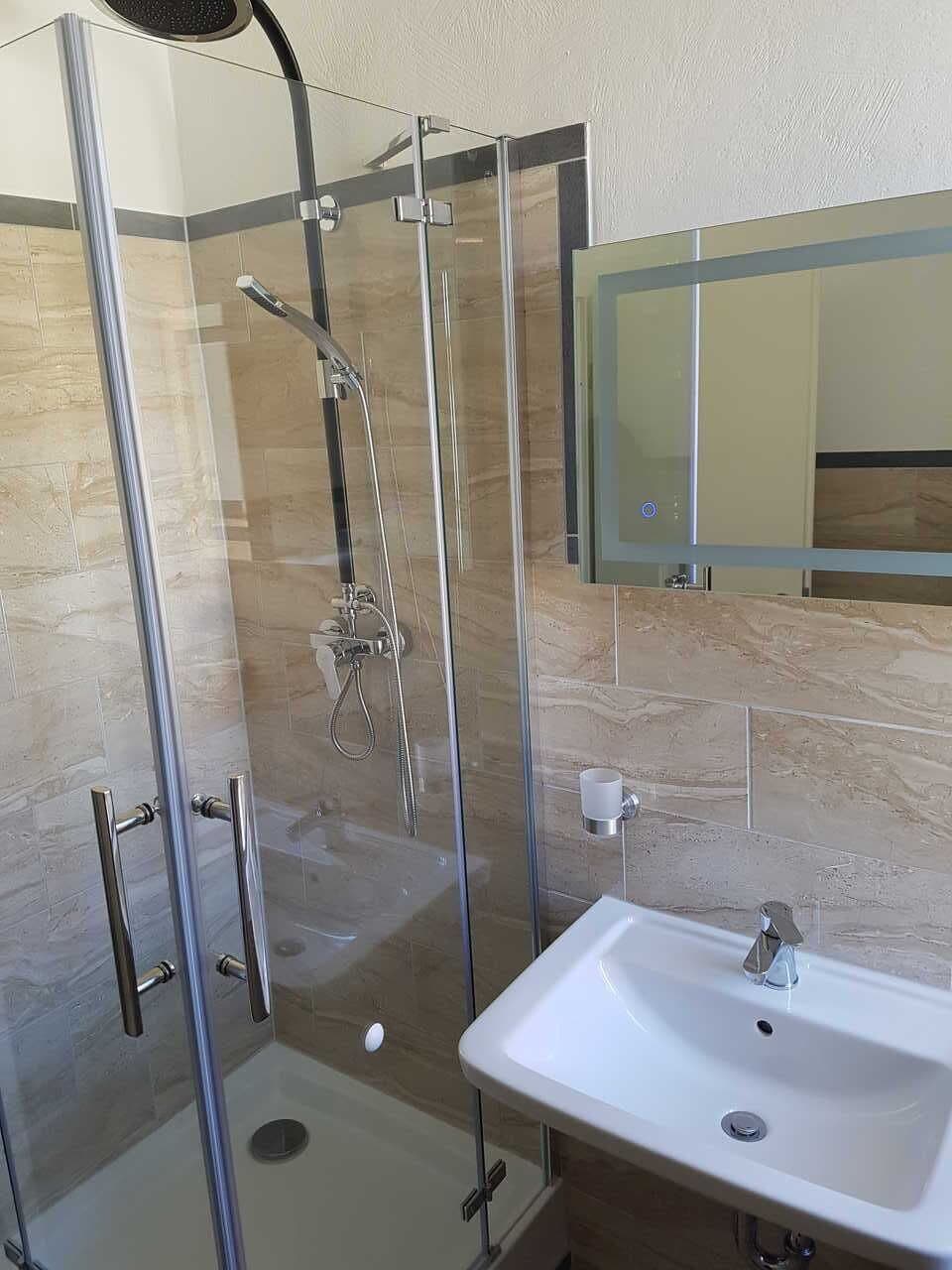 Egy modern fürőszoba, melyben üvegfalas zuhanyzó, fehér mosdókagyló és a mosdókagyló felett egy tükör található.